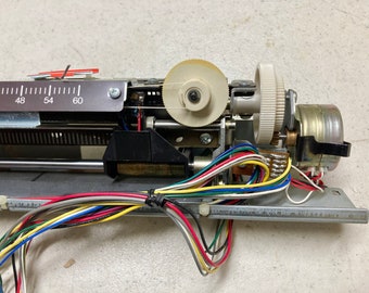 May include: Close-up of a mechanical device with gears, wires, and a numbered scale. The device has a silver metal frame, multiple colored wires, and a small motor. The scale reads from 42 to 60. The gears are white and beige.