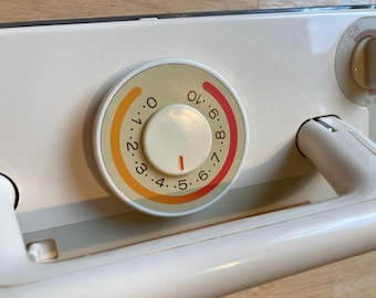 May include: Close-up of a cream-colored appliance control panel. A circular dial with numbers from 0 to 10 in orange and red, and a white center. A white handle and a knob labeled "CR" are also visible.