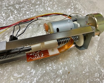 May include: Close-up of a mechanical device with a silver metal frame, a small motor, and a spring. Wires in red, yellow, and black are visible, along with a small circuit board and a white plastic component. The device appears to be electronic.