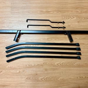 May include: Black metal bed frame components, including a long rectangular headboard support, curved side rails, and two smaller support pieces with hardware. The pieces are arranged on a wood-grain surface.