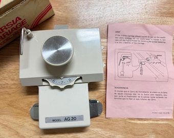 May include: A beige knitting machine carriage with a silver dial and a gauge marked with numbers. A pink instruction sheet and a cardboard box are also visible. The model number AG 20 is printed on the carriage.