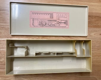 May include: An open, off-white plastic sewing kit with a removable lid. The lid features a pink illustration of sewing tools. The base of the kit has several compartments and shelves for organizing sewing supplies.