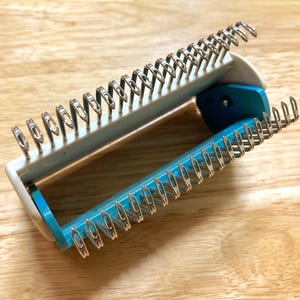 Peut inclure: Un outil de tricotage blanc et turquoise avec des boucles métalliques argentées. L'outil est ouvert, révélant l'intérieur, et repose sur une surface en bois brun clair. L'outil est conçu pour créer des articles tricotés.