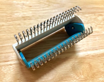 May include: A white and teal knitting tool with silver metal loops. The tool is open, revealing the interior, and sits on a light brown wooden surface. The tool is designed for creating knitted items.