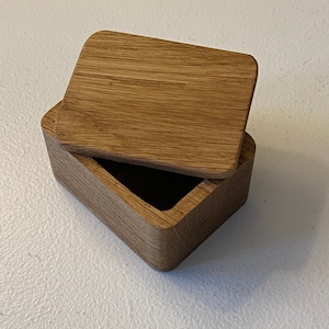 May include: A small wooden box with a hinged lid. The box is made of light brown wood and has a natural finish. The lid is slightly ajar, revealing the empty interior of the box.