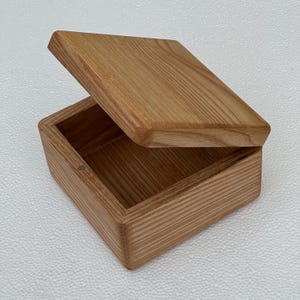 May include: A small, open wooden box with a hinged lid. The box is made of light-coloured wood with visible grain patterns. The lid is slightly ajar, revealing the interior. The box is square-shaped and appears to be handcrafted.