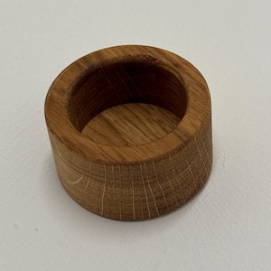 May include: A cylindrical wooden napkin ring with a natural wood grain finish. The ring is made of a light brown wood and has a smooth, rounded interior. The napkin ring is designed to hold a rolled napkin.