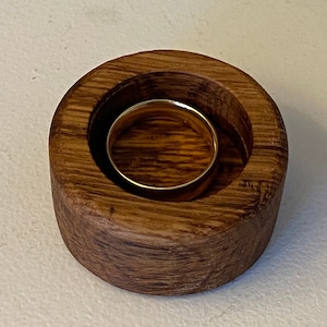 May include: A wooden ring holder with a round, deep well to hold a ring. The holder is made of dark brown wood with a natural grain pattern.