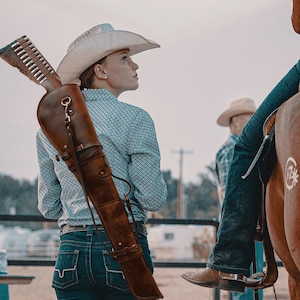 May include: A woman wearing a cowboy hat and blue jeans is carrying a brown leather rifle scabbard. The scabbard is strapped across her back and has a buckle. The woman is looking to the right of the image.