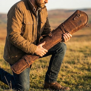 Pode incluir: Um estojo de rifle de couro marrom com uma alça e correias. O estojo é segurado por uma pessoa ajoelhada em um campo gramado. O estojo tem uma fivela e um anel em D. A pessoa está usando uma jaqueta marrom e jeans azuis.