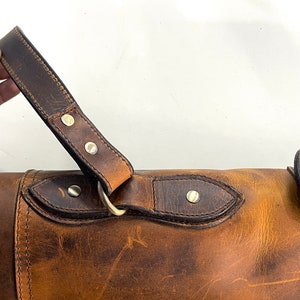 May include: A brown leather carrying case with a strap and a brass buckle. The case has a distressed look and is made of high-quality leather.