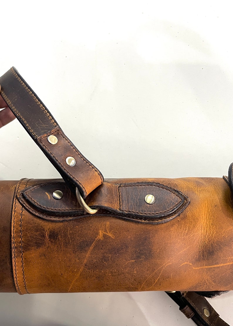 May include: A brown leather carrying case with a strap and a brass buckle. The case has a distressed look and is made of high-quality leather.