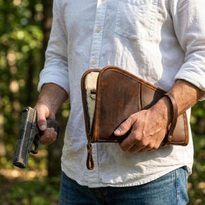 Pode incluir: Um estojo de couro castanho escuro com fecho de correr, forro interior cor de creme e alça de pulso, segurado ao lado de uma pistola semiautomática cinzenta. O estojo apresenta uma costura vertical e um puxador de fecho metálico.
