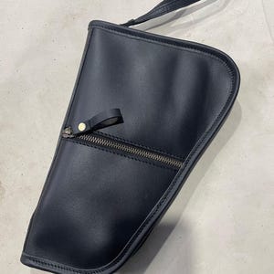Peut inclure: Une pochette de poignet en cuir noir avec une silhouette asymétrique et incurvée. La pochette présente une dragonne fixée sur le dessus, une poche zippée horizontale sur le devant avec une tirette en cuir et des coutures visibles sur les bords.