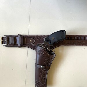 May include: A brown leather gun belt with a holster and a black revolver. The belt has a decorative floral pattern and a silver buckle. The holster is also decorated with a floral pattern.