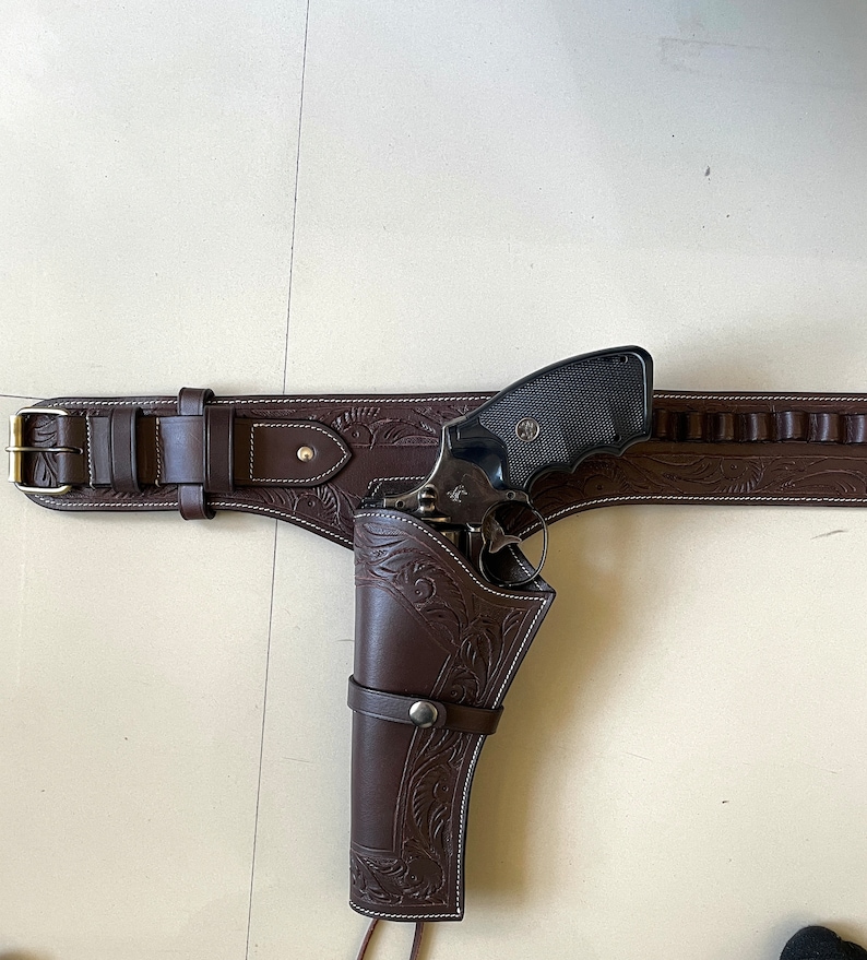 May include: A brown leather gun belt with a holster and a black revolver. The belt has a decorative floral pattern and a silver buckle. The holster is also decorated with a floral pattern.