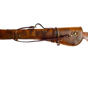 May include: A brown leather rifle scabbard with a strap and buckle. The scabbard is designed to carry a rifle and has a brass hook on the side.