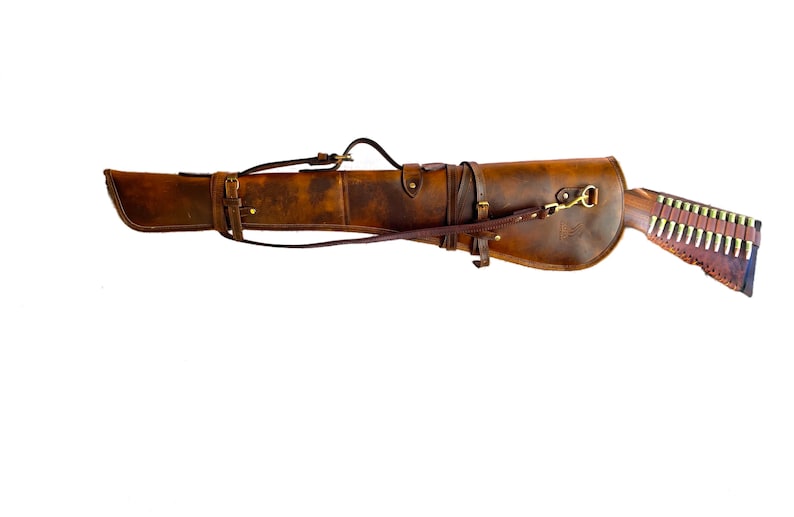 May include: A brown leather rifle scabbard with a strap and buckle. The scabbard is designed to carry a rifle and has a brass hook on the side.