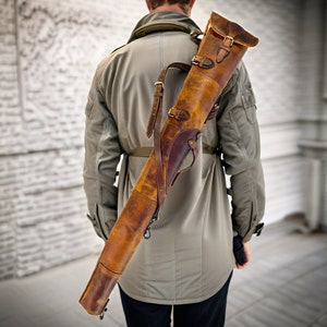 May include: A brown leather gun case with straps and buckles. The case is worn over the shoulder of a person wearing a beige jacket.