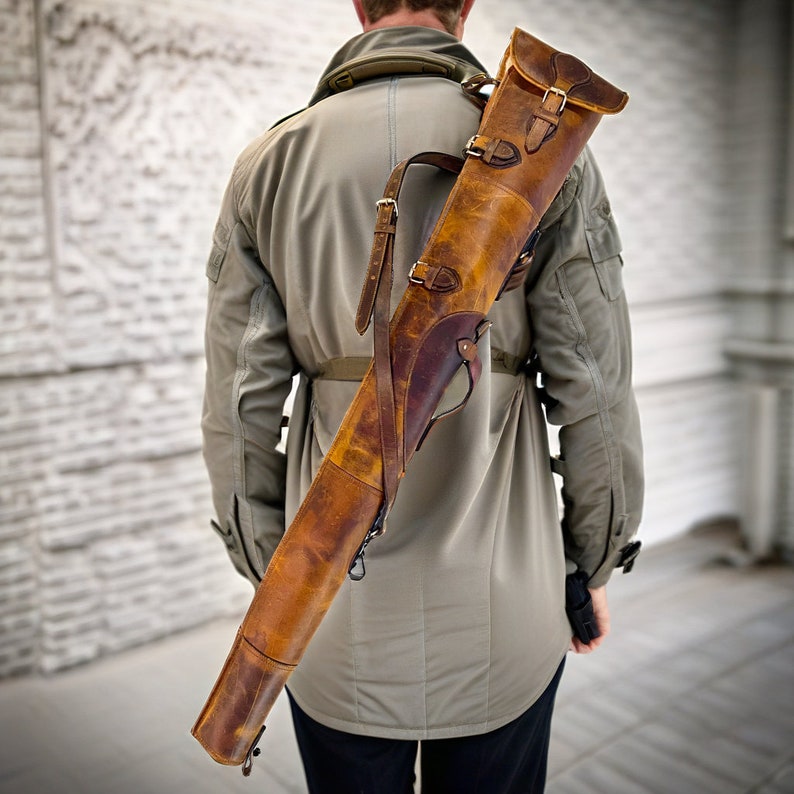 May include: A brown leather gun case with straps and buckles. The case is worn over the shoulder of a person wearing a beige jacket.