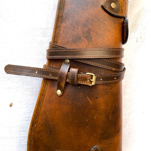 May include: A brown leather gun case with a strap and buckle. The case has a stitched design and a logo that says "ZOTO" with a flame design.