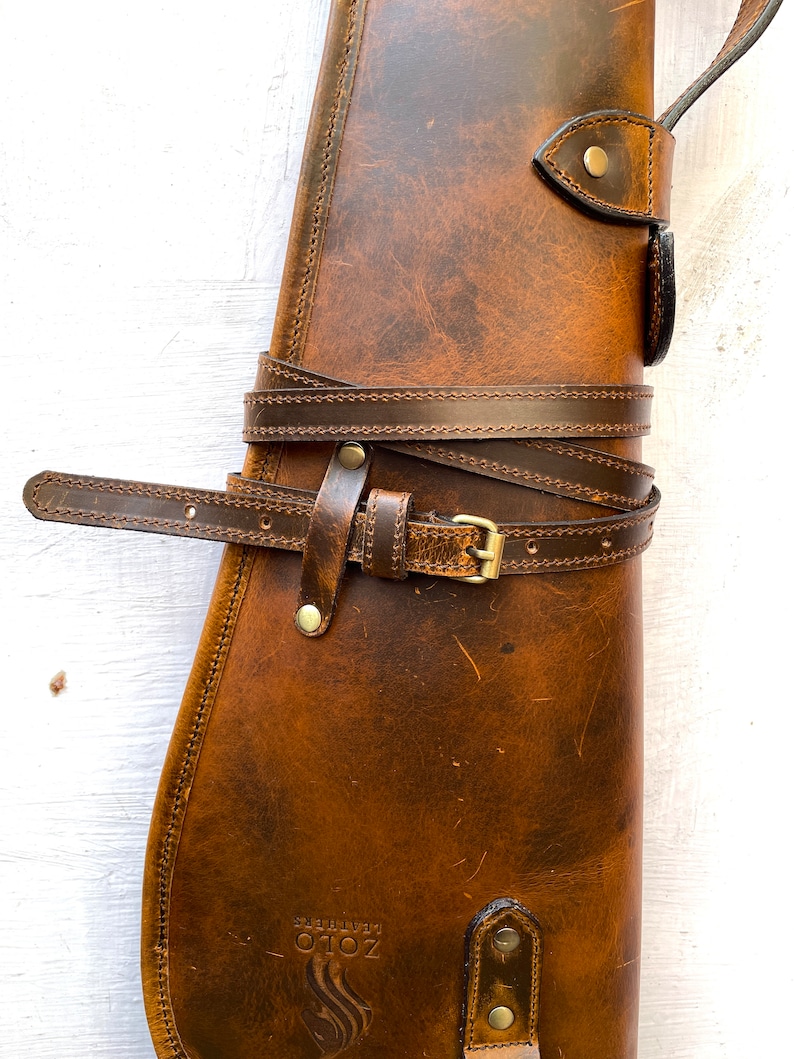 May include: A brown leather gun case with a strap and buckle. The case has a stitched design and a logo that says "ZOTO" with a flame design.