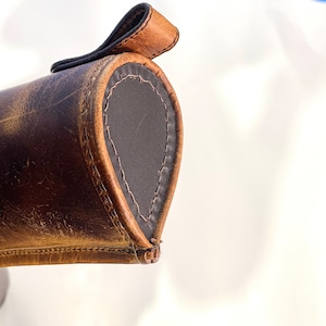 May include: Close-up of a brown leather case with a flap closure. The case has a dark brown leather lining and is stitched with brown thread.