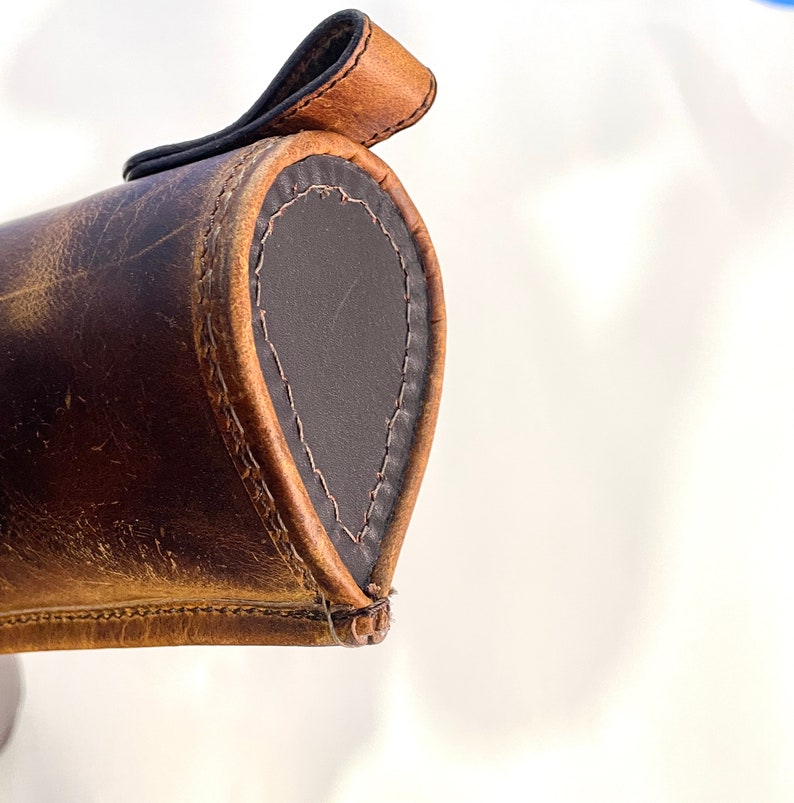 May include: Close-up of a brown leather case with a flap closure. The case has a dark brown leather lining and is stitched with brown thread.