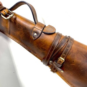 May include: A brown leather gun case with a strap and buckle. The case has a stitched design and a flap with a buckle.