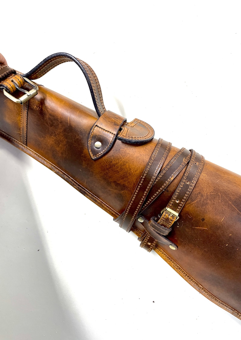 May include: A brown leather gun case with a strap and buckle. The case has a stitched design and a flap with a buckle.