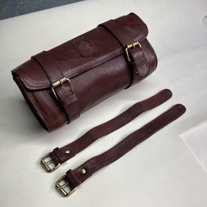 May include: A dark brown leather roll bag with two matching straps. The bag has a rectangular shape with a flap closure and two buckle straps. The straps have brass-colored buckles. A small embossed emblem is visible on the bag.