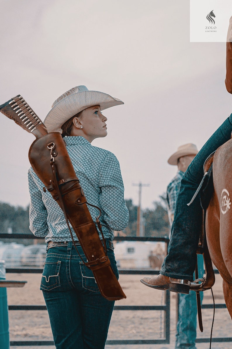 May include: A woman wearing a cowboy hat and blue jeans is carrying a brown leather rifle scabbard. The scabbard is strapped across her back and has a buckle. The woman is looking to the right of the image.