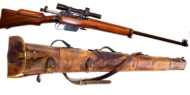 May include: A brown leather rifle case with a strap and buckles. The case is closed and has a dark brown leather patch on the top. A wooden rifle with a scope is shown above the case.