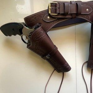May include: A brown leather gun belt with a silver buckle and two holsters. The holsters are decorated with an embossed floral pattern. The belt is tied with brown leather laces.