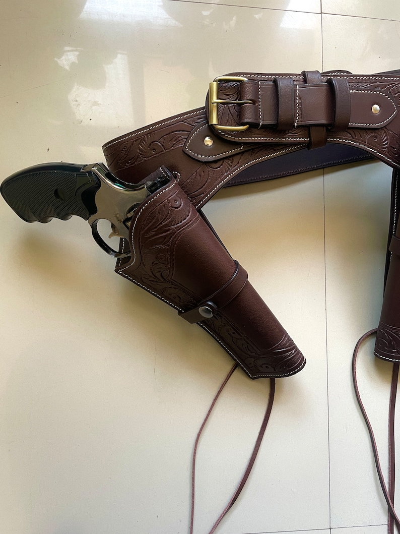 May include: A brown leather gun belt with a silver buckle and two holsters. The holsters are decorated with an embossed floral pattern. The belt is tied with brown leather laces.