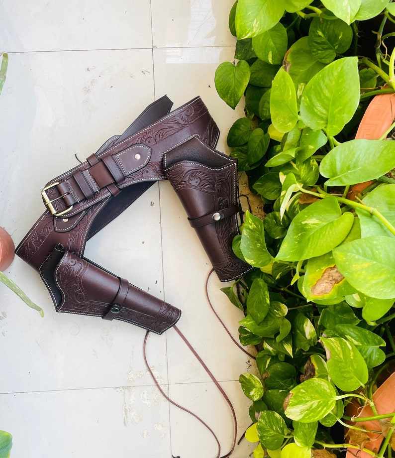 May include: A brown leather gun belt with two holsters. The belt has intricate floral designs and stitching. The holsters are also decorated with floral designs and have a strap to secure the gun.