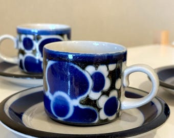 May include: Two ceramic espresso cups and saucers. The cups and saucers are decorated with a blue floral pattern on a white background. The cups have white handles and the saucers have a black and blue rim.