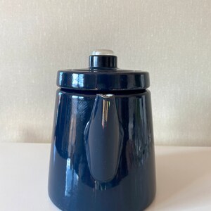 May include: A dark blue enamel teapot with a white knob on the lid. The teapot has a rounded body and a spout on the side.
