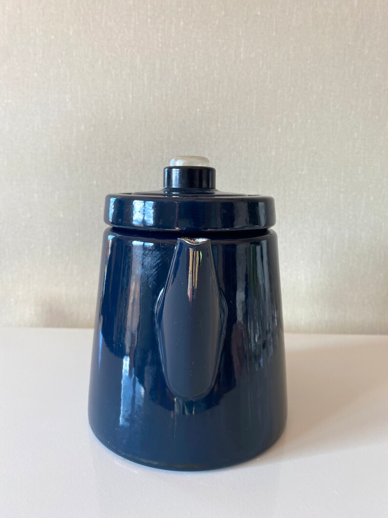 May include: A dark blue enamel teapot with a white knob on the lid. The teapot has a rounded body and a spout on the side.