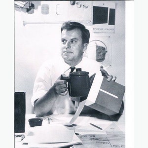 May include: A man in a white shirt sits at a desk holding a black and silver coffee pot. He is holding a white box with the text "FINLANDE - FRANCE" on it.