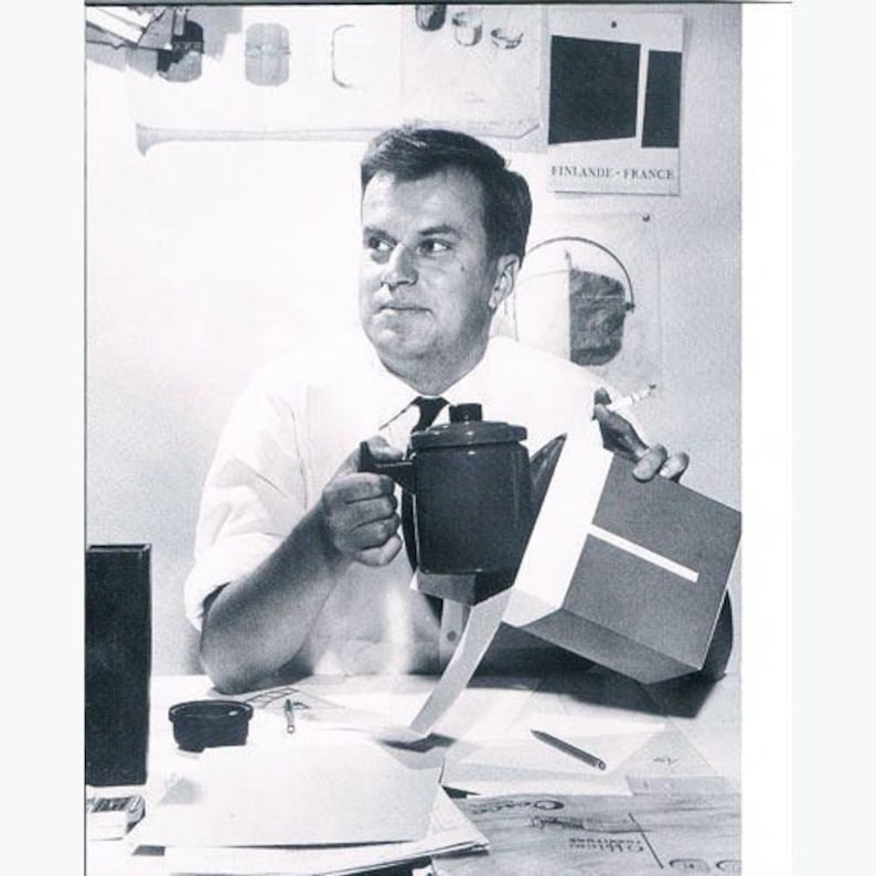 May include: A man in a white shirt sits at a desk holding a black and silver coffee pot. He is holding a white box with the text "FINLANDE - FRANCE" on it.