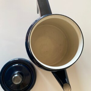 May include: A blue enamel teapot with a black handle and a white interior. The teapot has a spout and a lid. The lid is also blue enamel with a black knob.
