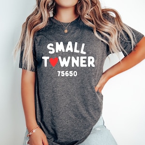 May include: A dark gray t-shirt with white text that reads "SMALL TOWNER" with a red heart in place of the "O". The number "75650" is printed below the text.