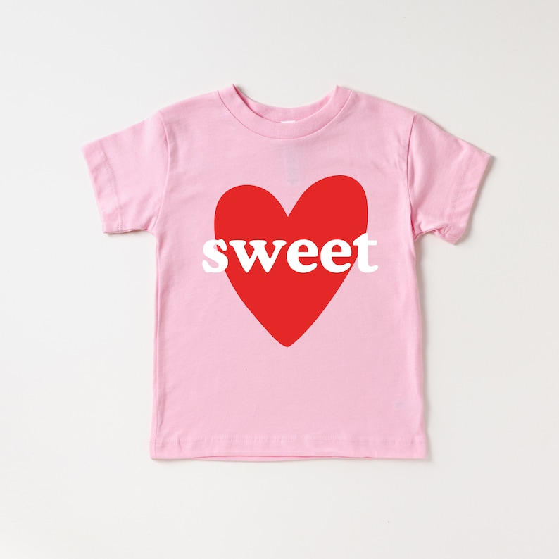 May include: A light pink t-shirt featuring a large red heart with the word "sweet" in white block letters. The shirt has short sleeves and a crew neck. The design is centered on the front of the shirt.