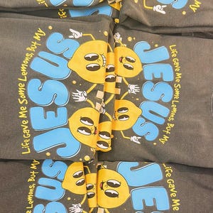 Op de afbeelding: Verschillende donkergrijze sweatshirts met het woord "JESUS" in blauwe letters en een cartoon citroen karakter. De tekst "Life Gave Me Some Lemons, But My" staat ook op de shirts. De citroenen zijn geel met zwarte details.