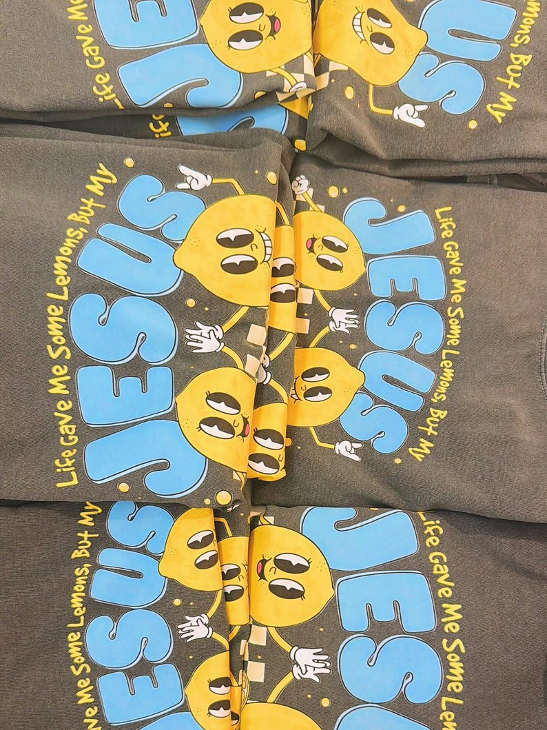 Op de afbeelding: Verschillende donkergrijze sweatshirts met het woord "JESUS" in blauwe letters en een cartoon citroen karakter. De tekst "Life Gave Me Some Lemons, But My" staat ook op de shirts. De citroenen zijn geel met zwarte details.