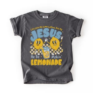 Op de afbeelding: Donkergrijs T-shirt met opgerolde mouwen met een retro-ontwerp. De afbeelding bevat twee cartoon citroenen, een glas limonade en de tekst "Life Gave Me Some Lemons, But My JESUS He Be Makin' LEMONADE."