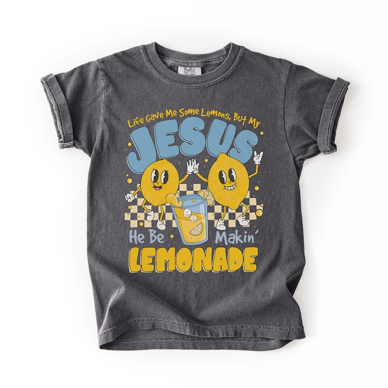 Op de afbeelding: Donkergrijs T-shirt met opgerolde mouwen met een retro-ontwerp. De afbeelding bevat twee cartoon citroenen, een glas limonade en de tekst "Life Gave Me Some Lemons, But My JESUS He Be Makin' LEMONADE."