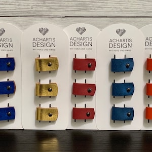 Könnte beinhalten: Fünf Karten mit dem Text "Achartis Design" und "Mit Herz und Hand" darauf. Jede Karte hat drei Lederriemen in verschiedenen Farben: blau, gold und rot.