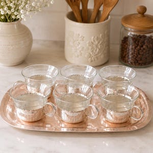 May include: A set of six clear glass cups with silver-toned metal bases featuring a decorative embossed pattern, arranged on a matching oval-shaped silver-toned metal tray with a textured surface and a slightly raised rim.